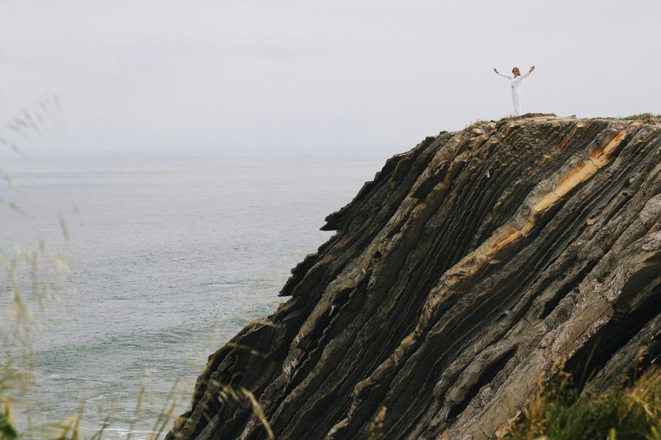 A lone woman in white stands triumphantly on a dramatic ocean cliff edge, embracing the vastness.
